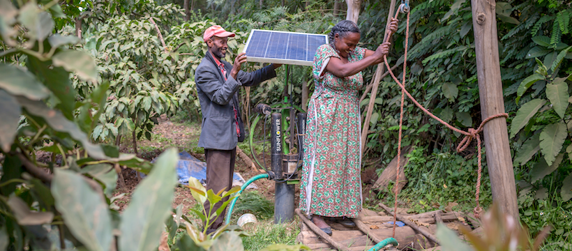 A women lowering down into a whole something on a rope, A man adjusting a solar panel on top of a small irrigation pump.