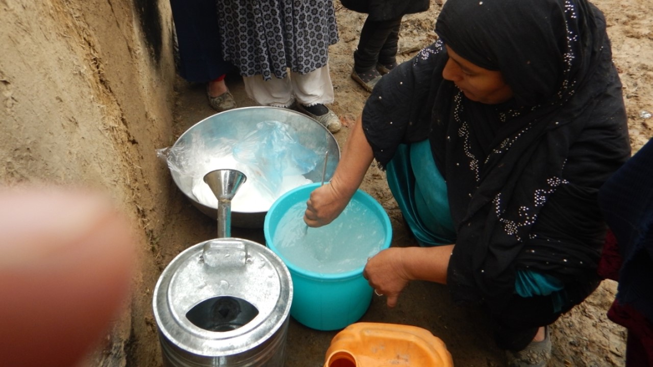 A woman in a headscarf mix somthing in a blue water container, with two metal can on either side of the water container.